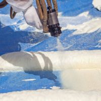 Technician dressed in a protective white uniform spraying foam insulation using Plural Component Spray Gun. Spraying polyurethane foam for roof and energy saving